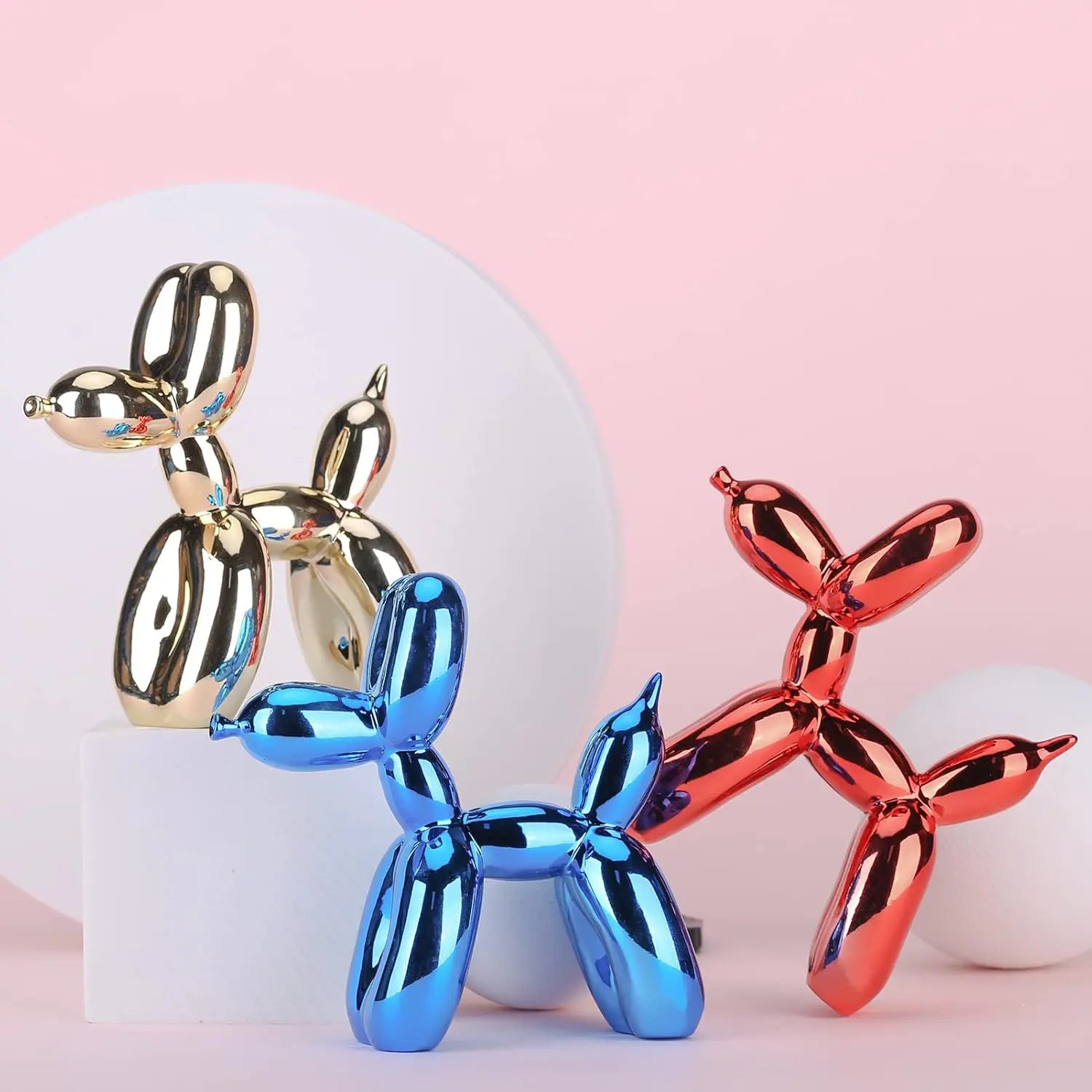 Gold, red, and blue metallic balloon dog sculptures arranged decoratively against a soft pink background with geometric props